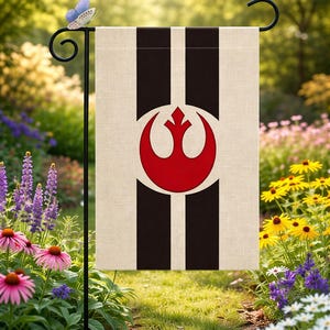 May include: A garden flag featuring a design with a red symbol within a white circle, set against a beige background with two vertical black stripes. The flag is displayed in a garden with colorful flowers.