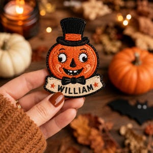 May include: An embroidered patch featuring a smiling orange jack-o'-lantern wearing a black top hat and a black bowtie. Below the pumpkin is a cream-colored banner with the name WILLIAM in black text, flanked by two small orange stars.