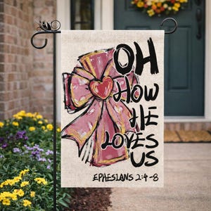 May include: A garden flag with the text "OH HOW HE LOVES US" and "EPHESIANS 2:4-8". The flag features a pink bow with a red heart in the center, set against a beige background. The flag is hanging on a black metal stand.