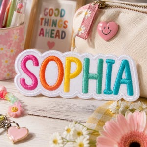 May include: A white embroidered patch featuring the name SOPHIA in colourful letters. The S is pink, O is orange, P is yellow, H is teal, I is teal, and A is teal. The patch has a scalloped white border and is placed in front of a canvas pouch.