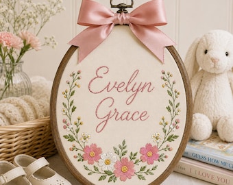 Peut inclure: Un tambour à broder ovale en bois contenant un tissu crème avec le prénom Evelyn Grace brodé en écriture rose, entouré d'une couronne de fleurs roses et blanches avec des feuilles vertes, surmonté d'un nœud en ruban de satin rose poudré.