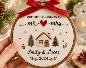 May include: A round wooden embroidery hoop containing off-white fabric with hand-stitched text reading OUR FIRST CHRISTMAS AS mr & mrs, Emily & Lucas, and 2024. The design features a house, two pine trees, stars, and a red ribbon bow on top.