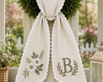 Peut inclure: Une décoration en forme de cœur en tissu rayé beige et blanc avec deux longs pans à bords festonnés. Un pan présente un motif floral brodé de marguerites blanches et de feuilles vertes, l'autre une grande lettre B verte avec des feuillages.