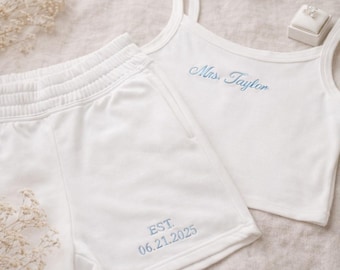 Peut inclure: Ensemble de détente de mariée blanc. Le débardeur est brodé de "Mrs. Taylor" en bleu clair, et le short porte la mention "EST. 06.21.2025". Un écrin à bagues est visible. L'ensemble est en tissu doux.
