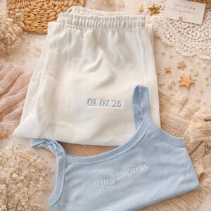 May include: A light blue vest top and white pajama bottoms are displayed. The vest top has the embroidered text "Mrs. Epifania", and the bottoms have the embroidered date "08.07.26". The sleepwear set is presented on a neutral surface.