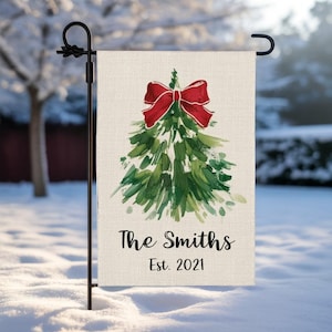 Puede incluir: Una bandera de jardín navideña personalizada con un árbol de Navidad en acuarela y un lazo rojo. La bandera muestra el nombre de la familia "The Smiths" y el año "Est. 2021". Se muestra en una estaca de jardín de metal negro.