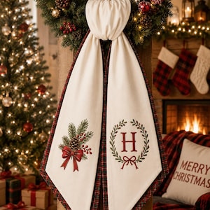 May include: A cream-coloured fabric runner featuring a pointed hem and a dark green and red plaid border. The left side displays an embroidered pinecone and berry motif with a red bow, while the right side features a red letter H inside a green wreath.