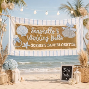 Op de afbeelding: Een rechthoekige stoffen banner met lichtblauwe verticale strepen aan de randen en een centraal beige paneel. De tekst luidt Seashells & Wedding Bells en SOPHIE'S BACHELORETTE, vergezeld door illustraties van een schelp, een ring en klokken.