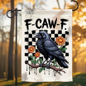 Könnte beinhalten: Eine Gartenflagge mit einem schwarzen Raben auf einem Ast, orangefarbenen Blumen und dem Text "F-CAW-F" vor einem karierten Hintergrund. Die Flagge wird im Freien an einem schwarzen Metallständer befestigt.