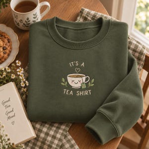 May include: A dark olive green long-sleeve sweatshirt featuring an embroidered graphic of a smiling teacup with a tea bag. The text above and below the cup reads, "IT'S A TEA SHIRT," with a small heart icon above the cup and leaves on the sides.