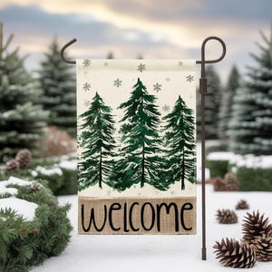 Peut inclure: Un drapeau de jardin avec une scène hivernale : trois pins verts et des flocons de neige. Le mot "WELCOME" est imprimé en noir sur une base texturée en toile de jute. Le drapeau est présenté sur un support en métal foncé, idéal pour la décoration extérieure.