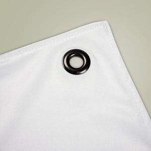 May include: Close-up of a white fabric corner with a dark gray grommet. The fabric has a subtle texture and is neatly stitched along the edges. The grommet is circular and has a shiny finish.