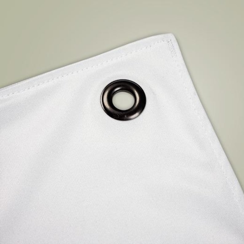 May include: Close-up of a white fabric corner with a dark gray grommet. The fabric has a subtle texture and is neatly stitched along the edges. The grommet is circular and has a shiny finish.
