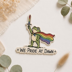Könnte beinhalten: Ein gestickter Aufnäher mit einem grünen Frosch, der einen Regenbogenumhang trägt und ein regenbogenfarbenes Schwert in die Höhe hält. Unter dem Frosch steht auf einem weißen Banner in schwarzer Schrift WE PRIDE AT DAWN, flankiert von zwei Sternen.
