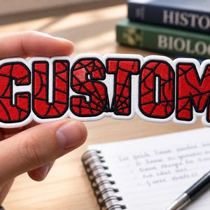 May include: A rectangular embroidered patch featuring the word CUSTOM in bold, red, block letters with a black spiderweb pattern overlay. The lettering is outlined in thick white thread, creating a distinct border around each individual character.