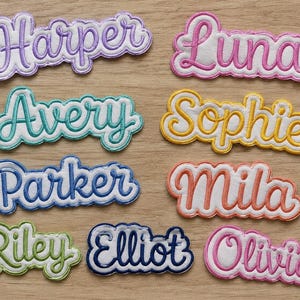 Könnte beinhalten: Eine Sammlung von neun bestickten Namensaufnähern mit gewelltem Rand und kursiver Schrift. Die Namen umfassen Harper in Lila, Luna in Pink, Avery in Türkis, Sophie in Gelb, Parker in Blau, Mila in Koralle, Riley in Grün, Elliot in Marineblau und Olivia in Magenta.