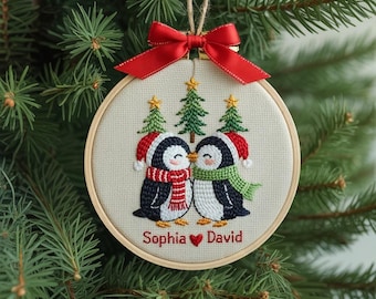 May include: Embroidered Christmas ornament featuring two kissing penguins wearing Santa hats and scarves. The ornament has a red bow and the names "Sophia" and "David" are stitched below the penguins. The ornament is displayed against a backdrop of evergreen branches.