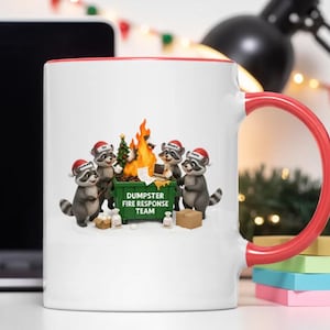 Pode incluir: Caneca de cerâmica branca com alça e borda vermelhas. A caneca apresenta uma ilustração humorística de guaxinins usando chapéus de Papai Noel em torno de um incêndio em uma lixeira. A lixeira diz "Dumpster Fire Response Team".