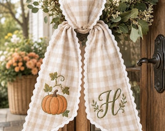 Peut inclure: Une décoration en forme de cœur en tissu vichy beige et blanc avec deux longs pans à bords festonnés. Un pan présente une citrouille orange brodée avec des feuilles vertes, et l'autre pan affiche une lettre H brodée en vert.