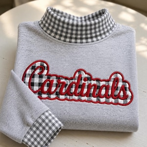 May include: A heather grey sweatshirt featuring a mock neck collar and sleeve cuffs in a black and white gingham pattern. The front displays the word Cardinals in red script lettering with a black and white gingham background and red trim.