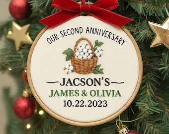 Peut inclure: Ornement de Noël brodé avec un cerceau en bois. Il présente un panier de coton, le texte « OUR SECOND ANNIVERSARY », les noms « JAMES & OLIVIA » et la date 10.22.2023. Un ruban rouge est attaché pour l'accrocher.