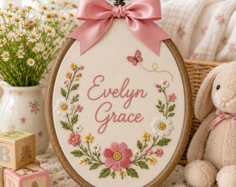 Peut inclure: Un tambour à broder ovale en bois avec le prénom Evelyn Grace brodé en fil rose cursif. Le motif comprend une couronne florale de fleurs roses, jaunes et blanches avec des feuilles vertes, un petit papillon rose et un ruban en satin rose.