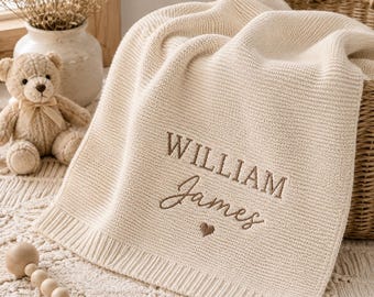 Peut inclure: Une couverture pour bébé en tricot couleur crème avec le prénom William James brodé en fil marron, agrémenté d'un petit cœur marron. La couverture présente une bordure côtelée et est présentée avec un ours en peluche et un hochet en bois.