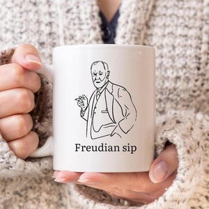 May include: White ceramic mug featuring a black line drawing of a man holding a cigar. The text "Freudian sip" is printed below the illustration. The mug is held by a person wearing a cream-colored sweater.