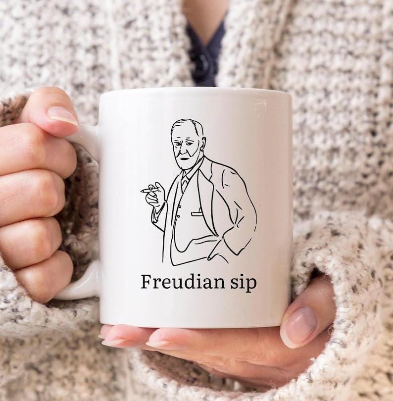 May include: White ceramic mug featuring a black line drawing of a man holding a cigar. The text "Freudian sip" is printed below the illustration. The mug is held by a person wearing a cream-colored sweater.