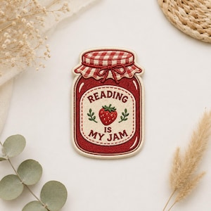 May include: An embroidered patch shaped like a mason jar filled with red jam. The jar features a red and white gingham lid and a cream-colored label with a red strawberry illustration and the text READING IS MY JAM in dark red lettering.