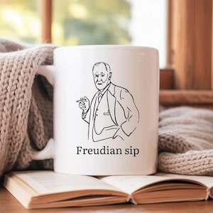May include: White ceramic mug featuring a black line drawing of a man in a suit, holding a cigar. The text "Freudian sip" is printed below the image. The mug is resting on an open book.