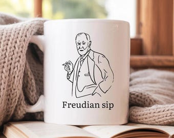 Peut inclure: Mug en céramique blanche avec un dessin au trait noir d'un homme en costume, tenant un cigare. Le texte "Freudian sip" est imprimé en dessous. Le mug est posé sur un livre ouvert.