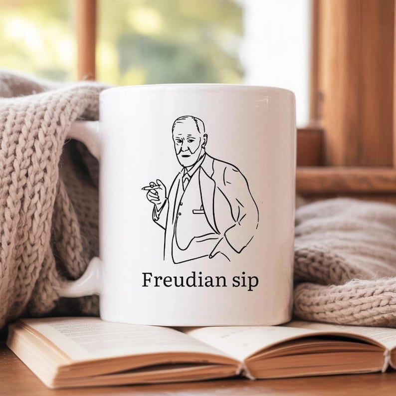 May include: White ceramic mug featuring a black line drawing of a man in a suit, holding a cigar. The text "Freudian sip" is printed below the image. The mug is resting on an open book.