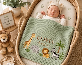 Peut inclure: Une couverture pour bébé en tricot vert sauge ornée d'animaux de safari brodés, dont une girafe, un éléphant, un lion et un zèbre, avec le nom OLIVIA rose cousu au centre. La couverture recouvre un bébé allongé dans un couffin en osier.