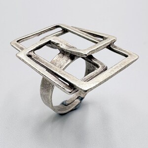 May include: A silver ring with a geometric design of overlapping squares. The ring has a textured, antique finish and a wide band. The openwork design gives the ring a modern, artistic look. The ring is set against a light grey background.