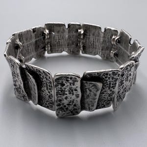Könnte beinhalten: Ein silberfarbenes Armband, das aus mehreren rechteckigen Gliedern mit einer stark texturierten, gehämmerten Oberfläche besteht. Jedes Glied weist in den Vertiefungen ein dunkles, oxidiertes Finish auf, was dem gesamten Stück ein robustes Aussehen verleiht.