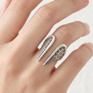 May include: A silver-toned open ring featuring two symmetrical wing-shaped components. Each wing element displays a detailed, intricate filigree pattern with textured cutouts, creating a delicate and airy appearance on the finger.