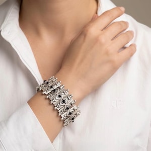 Könnte beinhalten: Ein breites Silberarmband mit einem sich wiederholenden geometrischen Muster aus rautenförmigen Gliedern. Jedes Glied ist in der Mitte mit einem kleinen, runden, polierten schwarzen Stein verziert und von winzigen Silberperlen umrandet.