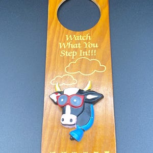 May include: Wooden door hanger with a cow design. The cow has red glasses and a blue bell. The text "Watch What You Step In!!!" is printed on the door hanger. The door hanger is brown with yellow accents.