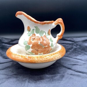May include: A small, white ceramic pitcher and basin set. The pitcher has a curved handle and spout, with orange and green floral designs. The rim of the pitcher and basin are trimmed in orange. The set is likely vintage.