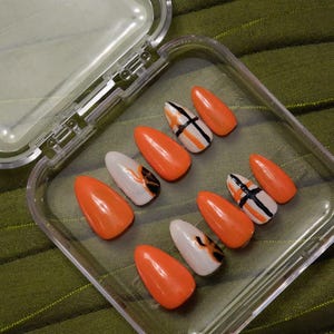 May include: A set of orange and white press-on nails in a clear plastic case. Some nails have black and white cross designs, while others feature a flame-like pattern. The nails are glossy and almond-shaped.
