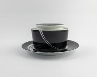 Puede incluir: Un cuenco de cerámica negra con tapa sobre un plato a juego. El cuenco y el plato tienen un interior blanco y un patrón decorativo de finas líneas blancas curvas que recorren las superficies exteriores de color negro brillante.