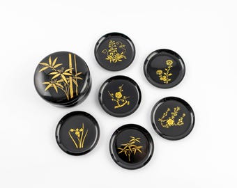 Puede incluir: Un juego de laca negra compuesto por un recipiente con tapa y cinco platos redondos pequeños. Cada pieza presenta ilustraciones botánicas doradas, que incluyen bambú, flores de ciruelo y varios motivos florales en las superficies.