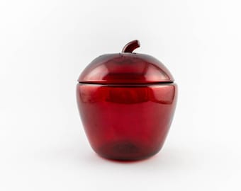 Puede incluir: Un tarro de cristal translúcido de color rojo intenso con forma de manzana. El recipiente presenta un cuerpo redondeado y una tapa extraíble a juego con un pequeño tallo curvado como asa en la parte superior. El cristal es liso.