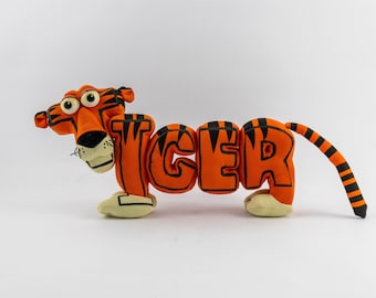 Puede incluir: Un peluche con forma de tigre de cuerpo naranja y rayas negras. El cuerpo está formado por las letras T, I, G, E y R, con la cabeza unida a la letra T y una larga cola rayada que sale de la letra R. Tiene patas de color blanco.