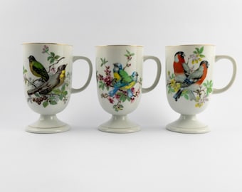 Puede incluir: Tres tazas de cerámica blanca con base de pedestal y bordes dorados. Cada taza presenta una ilustración detallada de aves posadas en ramas florecidas, mostrando varias especies con plumaje vibrante en verde, amarillo, azul y rojo.