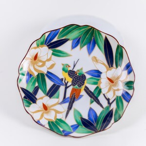 May include: White ceramic plate with a scalloped edge, featuring a colorful design of birds, white flowers with gold accents, and green and blue leaves. The plate has a brown rim.