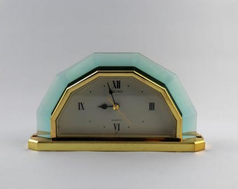 Puede incluir: Un reloj de sobremesa de cuarzo Seiko con un marco de cristal esmerilado azul claro semicircular y una base de metal dorado. La esfera blanca muestra números romanos negros en las posiciones de las tres, seis, nueve y doce.
