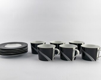 Puede incluir: Un juego de cuatro tazas de cerámica negra y cinco platos a juego. Cada taza presenta un interior blanco y un exterior negro con tres finas rayas blancas diagonales. Los platos son negros con un centro blanco y una línea circular.