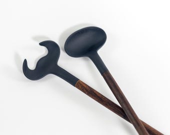 Puede incluir: Un par de utensilios de cocina negros con mangos de madera oscura. Uno es un cucharón, el otro un diseño en forma de tenedor. Los utensilios están angulados uno sobre otro sobre un fondo blanco. Cocina y comedor.